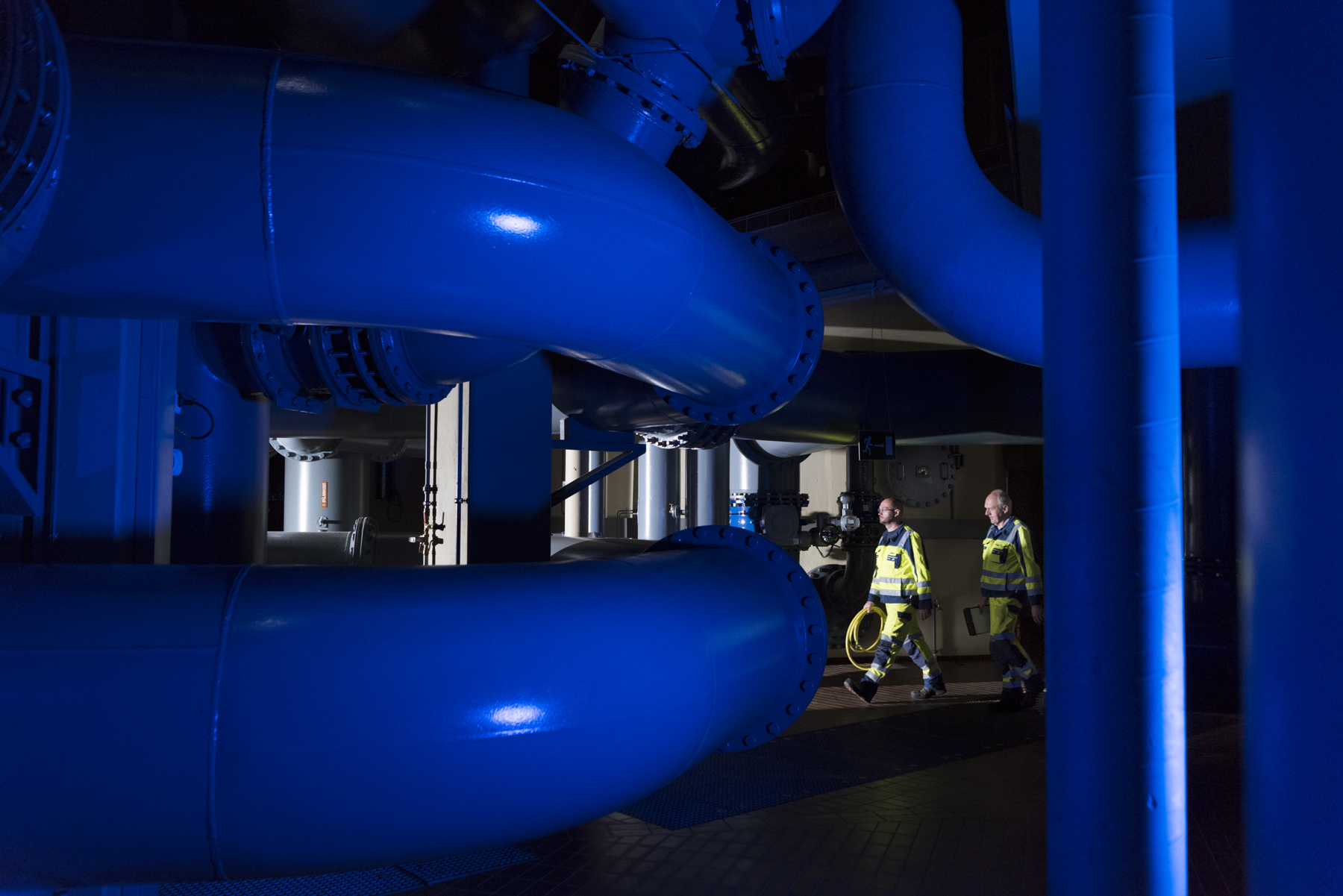 Blick ins Innere des Wasserwerkes Zwei Mitarbeiter gehen durch den unterirdischen Bereich des Wasserwerkes Panzenberg. Riesige, blaue Rohre sind im Vordergrund zu sehen.